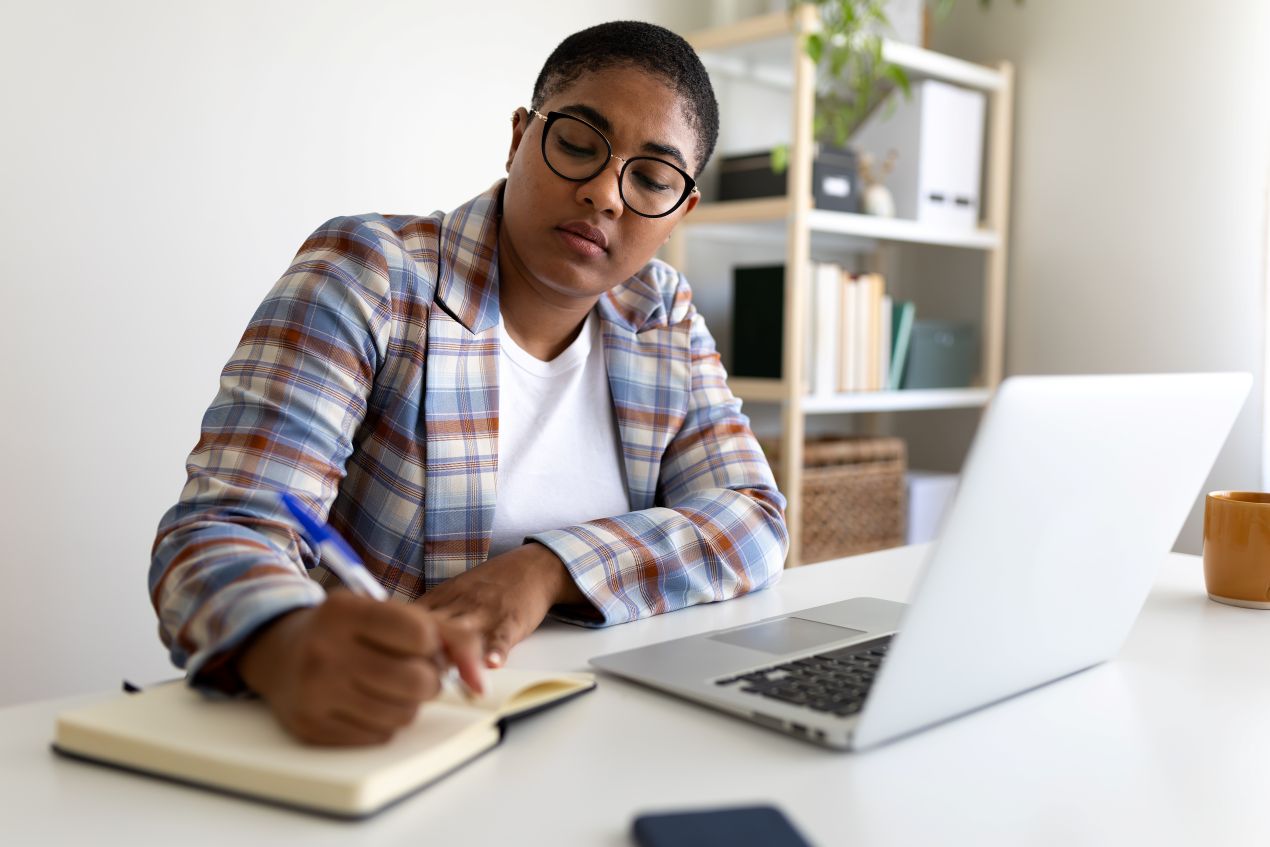 Frau mit Brille und kariertem Blazer schreibt Notizen am Laptop – organisation und textarbeit fuer ghostwriter hausarbeit im homeoffice.