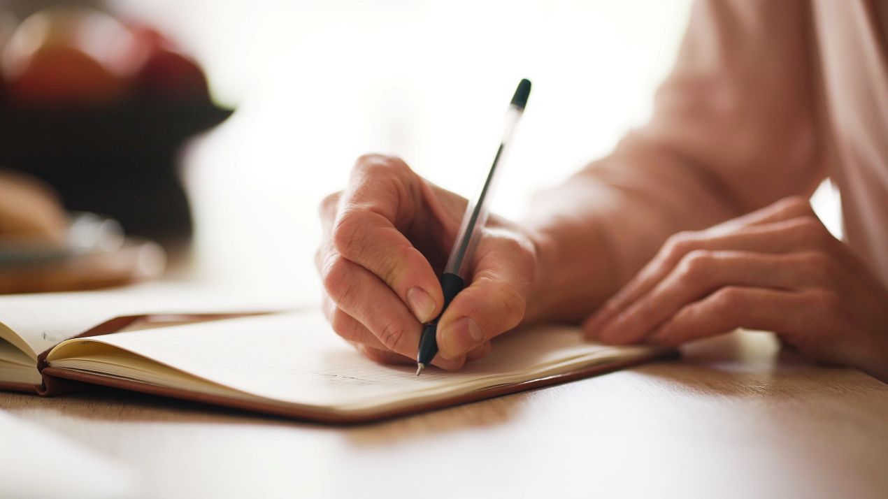 Person schreibt mit Stift in Notizbuch auf Holztisch – konzentriertes Arbeiten fuer ghostwriter hausarbeit, Planung und Textstruktur.