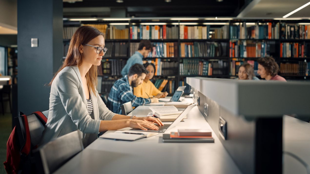 Studierende sitzt mit Laptop in Bibliothek und arbeitet fokussiert – inspiration und effiziente Arbeitsweise fuer ghostwriter hausarbeit.